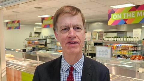 A man in a white and blue chequered shirt, red tie and blue suit jacket. He is looking into the camera and stood in front of a school canteen