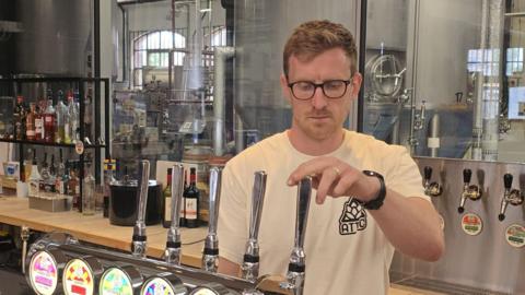 A man with blond hair stands behind a bar with his hand on a beer pump.