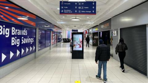 People walking past advertisement inside shopping centre which reads "big brands, big savings" 