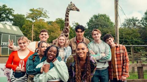 The cast of Heartstopper pose happily for a photo with a giraffe looming behind them