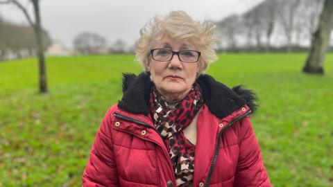 Pauline Lancaster in centre of the photo. She is wearing a red puffer coat with a black fur trim on the hood, with a matching red, brown and black scarf. Pauline is wearing lipstick and black glasses, her hair is blonde in a pixie cut. Pauline is stood in Cambridge Park. The grass is vibrant green and the trees are bare. It's glum, foggy and grey. 