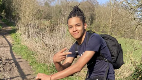 16-year-old Sean poses for the camera riding a bicycle on a country path. He has dreadlocked hair tied on the top of his head, and is wearing a blue t shirt and black backpack.