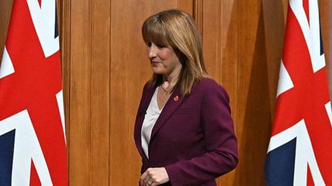 Rachel Reeves in a purple suit and white shirt. A Union Jack flag can be seen to her left and right