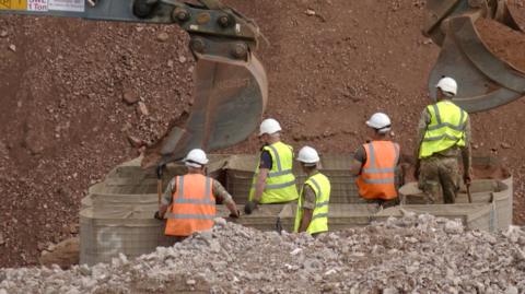 Five people, two in orange hi-vis vests and three in yellow ones, stand around a barrier constructed around an unseen 50kg bomb discovered in Coventry, while two diggers cover the device with sand to minimise damage during an explosion.