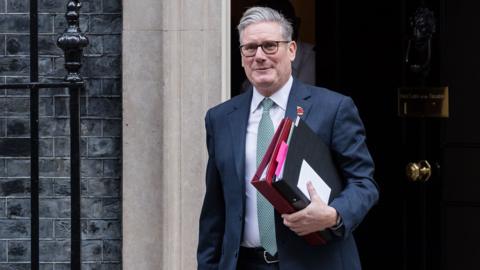 Keir Starmer leaves Downing Street while holding folders and wearing glasses and a suit and smiling slightly on Wednesday