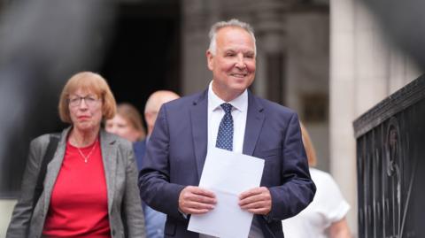 Chris Whitbread walking while smiling and clutching a piece of A4 paper. He has grey hair and is wearing a blue suit jacket over a white shirt, with a blue tie that includes white dots. To his right, slightly in the background, is a woman with brown hair and glasses and she is wearing a red top and grey blazer - she is the monitoring officer of Epping Forest District Council.