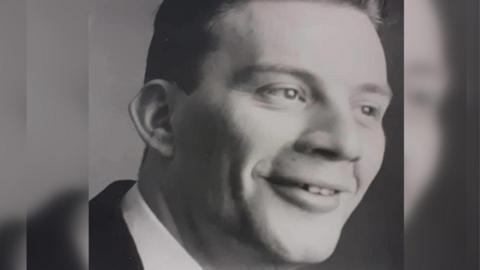 A black and white photo of Anthony Scarrott. He is facing sideways and is wearing a suit and a shirt.