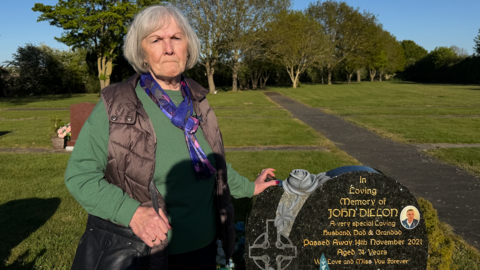 Ann Dillon is wearing a green jumper and a black jacket on top. She is standing next to her husband's grave, marked with a black and gold headstone. She has her left hand resting on it. 