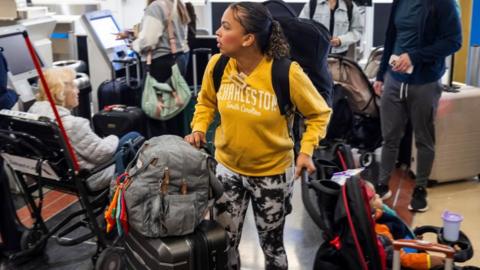 A woman wearing a backpack in an airport hangs onto a stroller with hand and a suitcase with the other