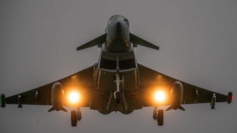 RAF Eurofighter Typhoon jet landing at RAF Coningsby after a sortie. The shot is taken from underneath and the aircraft has its landing gear down, with missile casings visible on the undercarriage.