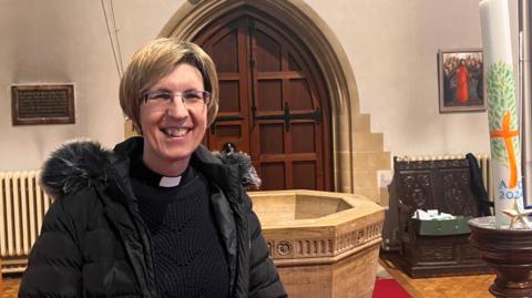 Reverend Robyn Golden-Hann is wearing a black jumper, a black shirt with white dog collar, and a black coat. She has glasses and medium length brown hair, and is stood in a church.