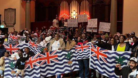 A group of about 100 people gathered in a hall with the people at the front of the group holding large flags of the Chagossians. They are standing in front of a large pipe organ.