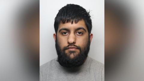 A police mugshot of Abdul Sami. He has black hair and a black beard. He also has brown eyes and is staring directly at the camera.