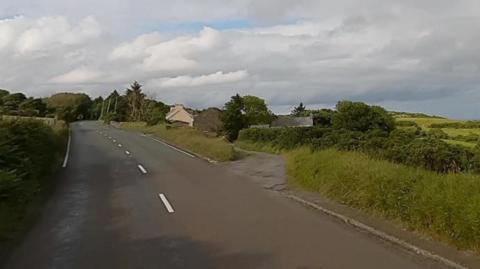 The coast road pointing in the direction of Ramsey. The tarmacked road, which has a broken white line painted down the middle and single white lines along each side, disappears off into in the distance, curving past a group of trees. There are green fields on both sides with a farm house and farm buildings visible on the right.