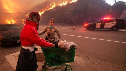 A police officer escorts a homeless woman to evacuate as fire rages down the hills in Pacific Palisades, on 7 January 7, 2025