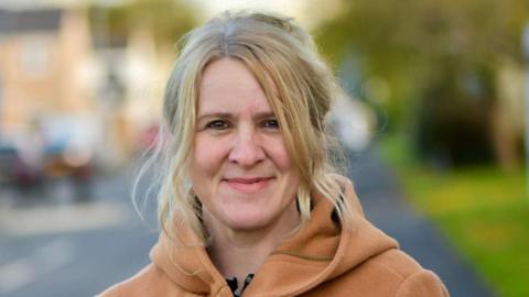 Councillor Ella Billiald a woman with wavy blonde hair half pulled up and an orangey-beige coat with a hood, her background is blurred