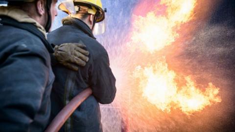 Two firefighters wearing dark uniforms and yellow helmets use a hose to spray water onto flames