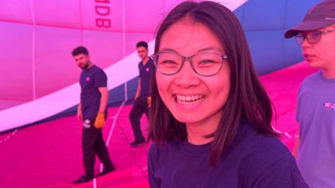 A woman smiling at the caera inside of a pink and blue hot air balloon. Behind her, there are three men.