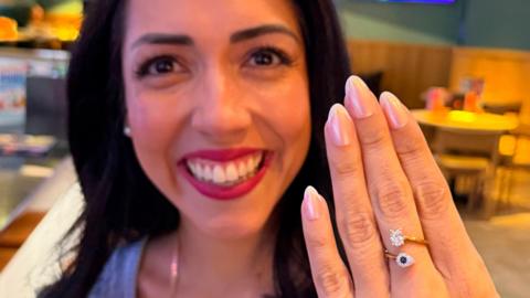 A close up of a young woman smiling broadly and holding her hand up in front of her. She has a ring on her middle finger with a sapphire and diamond.