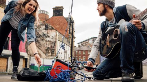 Busker with a black guitar crouching by an amp whilst a lady in a denim jacket gives money