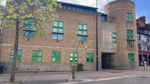 Luton Crown Court building entrance. A tree with green leaves stands in front of the the brown brick building with green windows 