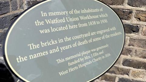 A close up of the grey, circular plaque that remembers the people that were housed at the workhouse. It says it's funded by a former patient of Watford General, Mrs J Ellis. 