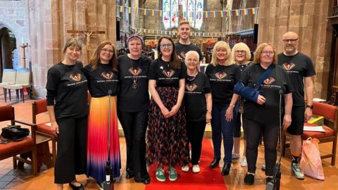 A group of people are inside a church. They are dressed in black T-shirts with a rainbow and a cross on them. Behind them, rainbow flags are strung up on banners.