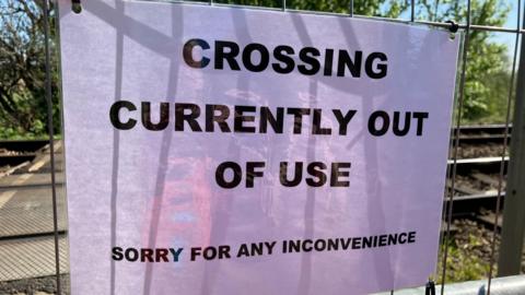 Notice on wire fence with railway lines behind saying CROSSING CURRENTLY OUT OF USE SORRY FOR ANY INCONVENIENCE