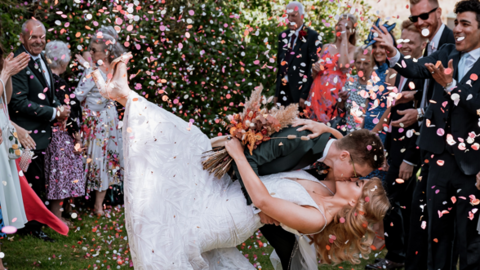 A man wearing a dark green wedding suit is kissing a woman with blonde hair on the lips and she is wearing a white lace wedding dress. There are people smiling and throwing confetti in the background 