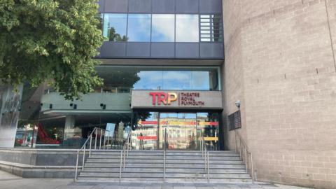 The exterior of the Theatre Royal Plymouth. There are steps leading up to the buildings which has glass doors which are reflecting the building opposite. There is a tree to the side.