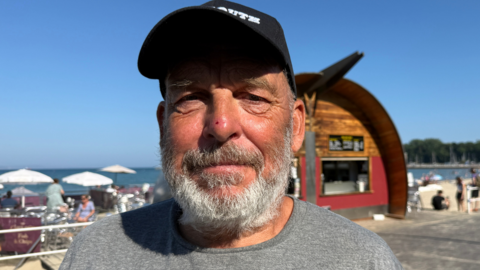 A man wearing a cap and a grey t-shirt looking into the camera on a sunny day next to the beach