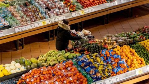 A woman in a dark coat and seen from above pushes a shopping cart through the produce section of a grocery store. On either side of her are piles of brightly coloured produce.