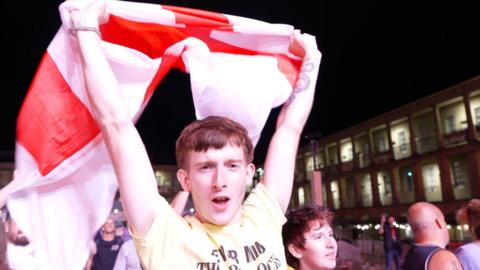 A man in a yellow t-shirt holds an red and white England flag aloft