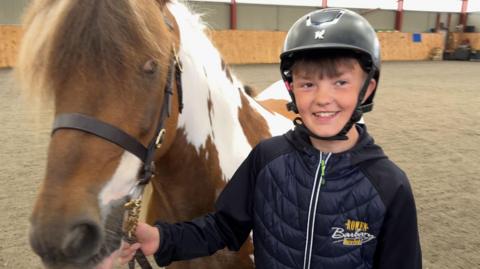 Theo stands on the right of the picture, holding Jerry, a brown and white horse. Theo is leading him with a black bridle and lead. Theo is wearing a black riding hat, and blue padded coat. He is smiling. He has brown eyes and under his hat you can see a dark brown fringe. 