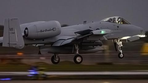 Grey coloured jet aircraft. The pilot can be seen in the cockpit. The wheels are down as it comes in to land. The background is blurred owing to the motion of the aircraft. The picture was taken at night.