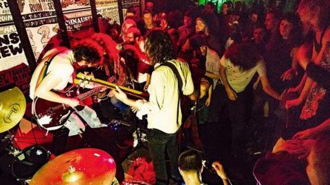 Two men hold guitars in a band with a crowd inside Mother's Ruin, Bristol.