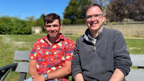 Adam, wearing a red shirt with flowers on sat on a bench next to David who is wearing a grey fleece. Both are looking at the camera and smiling. 