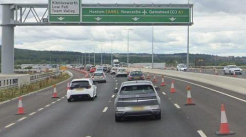 The A1 near Gateshead. There are four lanes but the two lanes at the edges are coned off, with cars going along the middle two lanes. A large gantry stands above the road with place names on it.