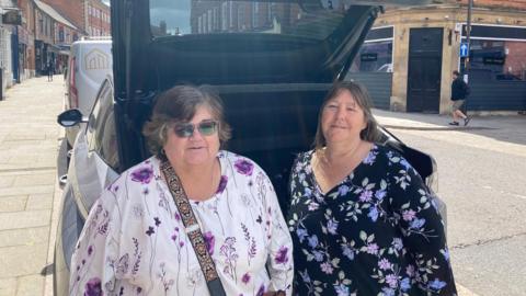 Two women wearing flowery tops propped against the back of a car with the boot open in an on-street bay in a street in a town centre with red-brick buildings to either side. The woman on the left has wavy dark hair and wears sunglasses, a white top with purple pattern and a dark shoulder strap. The woman on the right has dark, chin-length hair and wears a black top with pink and blue pattern.