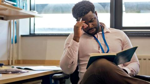 A teacher sits in a school office, reading from an open folder with his hand to his head. The desk nearby is scattered with papers and office supplies, and shelves above hold neatly arranged binders. There are windows behind the teacher letting in light.