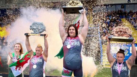 four strong women on a podium wearing gymnastics type body suits  with three holding trophies above their heads, including Rebecca in the middle with a welsh flag on her thigh and Sam with a Welsh flag draped on her shoulders. 