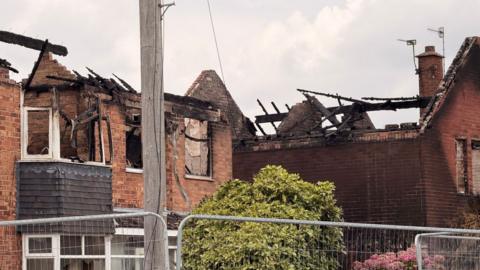 Two houses, with collapsed roofs, can be seen behind metal fencing. There are gardens in the foreground and a telegraph pole.