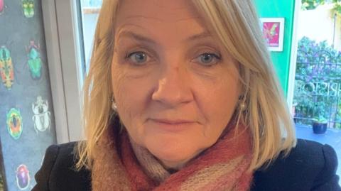 A selfie of Denise, who has shoulder-length blonde hair and blue eyes. She wears a pink and white striped scarf and dark jacket.