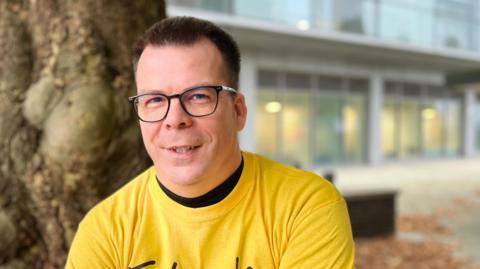 Aaron is wearing a yellow t-shirt. He has short brown hair and has black-rimmed glasses on. He is sitting outside under a tree with an office building blurred in the background.