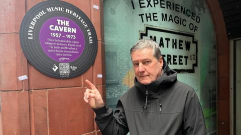 Peter Hooton is dressed in a black jacket and is standing while pointing at a heritage trail plaque which reads "The Cavern".