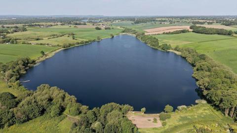 Aerial shot of Pickmere Lake in Cheshire on a sunny day, a wide body of water surrounded by green spaces.