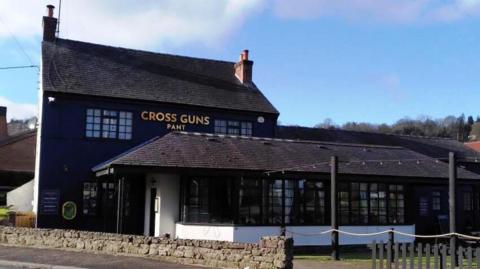 A large dark building with a dark tiled roof and an extension at the front with the words Cross Guns Pant in gold lettering on the wall