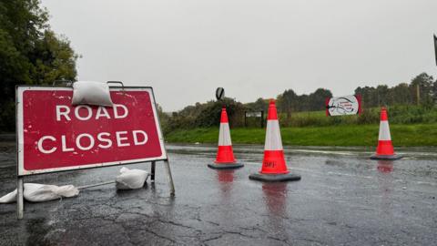 Road closed sign on a road with three white and orange cones. 