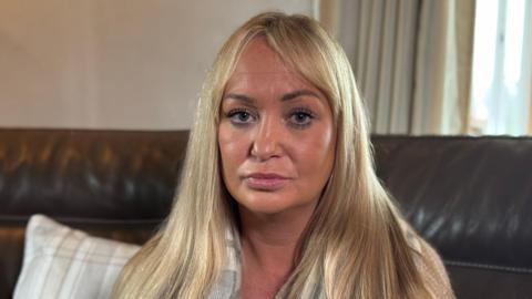 A woman with straightened blonde hair and makeup stares into the camera. She is sitting on a brown leather sofa