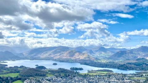 Grey and white clouds in blue sky over a stretch of blueish mountains, a loch and a small town